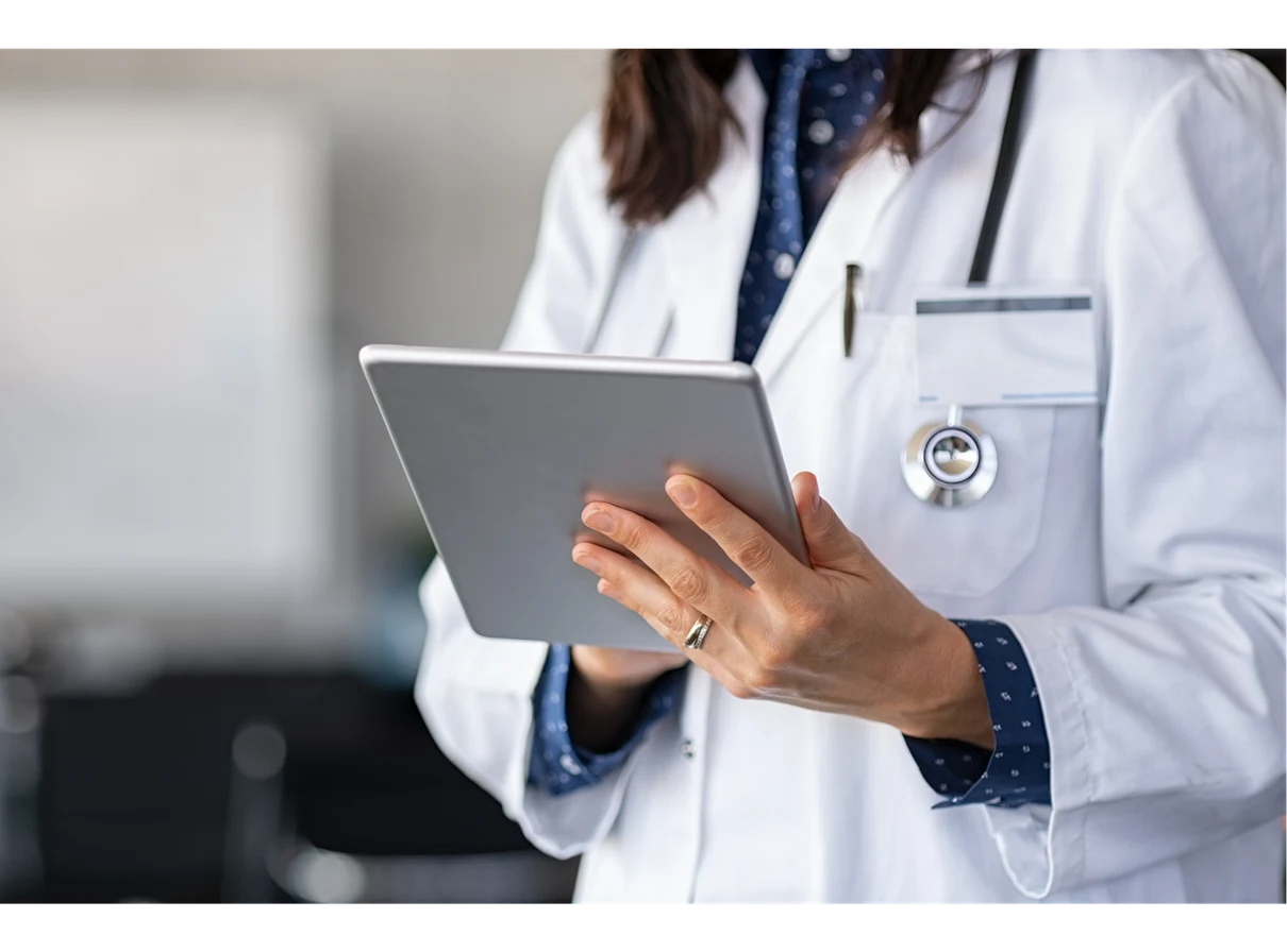 Doctor in a white lab coat holding and using a tablet, with a stethoscope around their neck and an ID badge visible.
